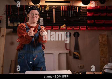 La donna falegname indossa abiti da lavoro nella scena dell'officina con attrezzi appesi al muro che fanno soldi. falegname donna in tarda serata in garage autentico. Foto Stock