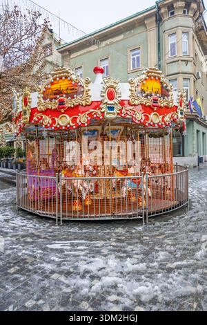 Una giostra natalizia si trova in una piazza urbana ricoperta di gelo, con cornici dorate, pannelli illuminati e cavalli da equitazione. I visitatori apprezzano un classico holi Foto Stock