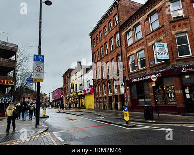 Manchester, Regno Unito - 21 gennaio 2026: Vivace Manchester Street con facciate in mattoni, cartelli e pedoni dopo la pioggia. Foto Stock
