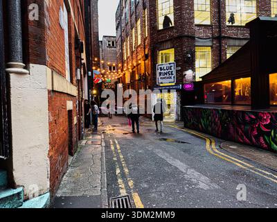 Manchester, Regno Unito - 21 gennaio 2026: Vicolo stretto di Manchester con pareti in mattoni, vetrine luminose e pedoni. Foto Stock