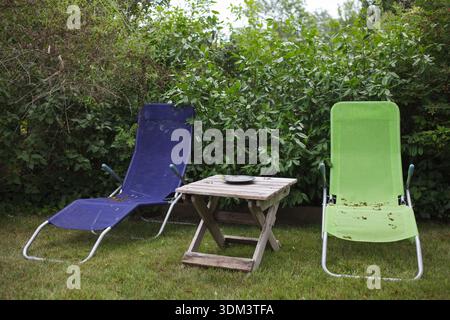 Due sedie da prato a rete colorata e un piccolo tavolo pieghevole in legno adagiato su una siepe verde verdeggiante a North Truro, Cape Cod. Foto Stock