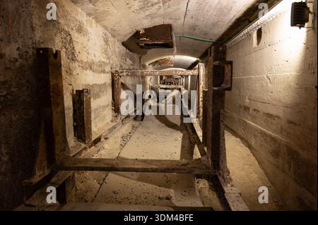 Vista interna profonda di un tunnel scuro di una fabbrica di cemento abbandonato, con supporti per nastri trasportatori arrugginiti e pareti in cemento polveroso Foto Stock