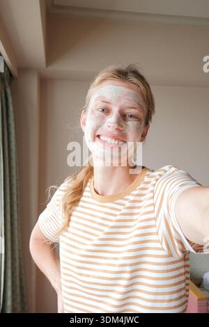 Donna con maschera cosmetica sul viso che si fa un selfie e sorride felicemente in camera d'albergo, idratando la pelle dopo il viaggio in aereo. Cura della pelle in viaggio || modello rilasciato Foto Stock