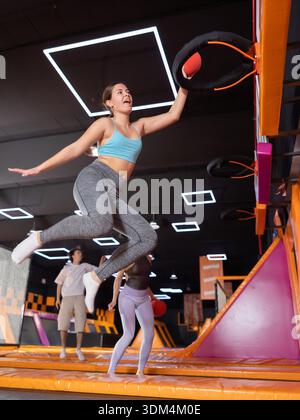 Giovane donna eccitata che ama divertirsi saltando in alto sul trampolino e colpendo il canestro da basket con la palla nel club di gioco Foto Stock