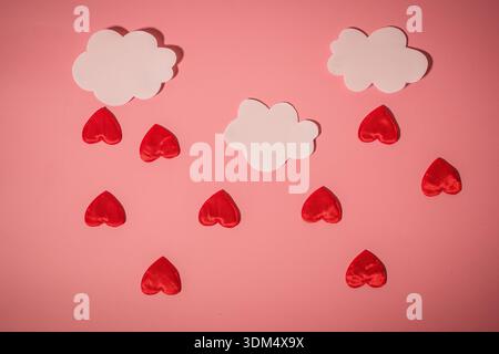 Due nuvole bianche sono appese sopra le forme rosse del cuore su uno sfondo rosa. Questa configurazione è stata creata per la festa di San Valentino. I cuori sono sparsi uniformemente Foto Stock
