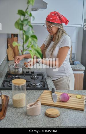 Donna incinta che cucina le uova in una pentola sul piano cottura, preparando un pasto sano a casa Foto Stock