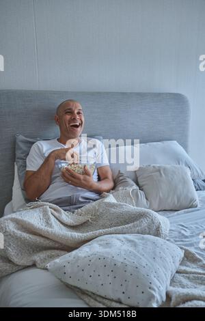 Uomo che ride tenendo in mano una ciotola di popcorn godendosi l'intrattenimento a letto la sera del film Foto Stock