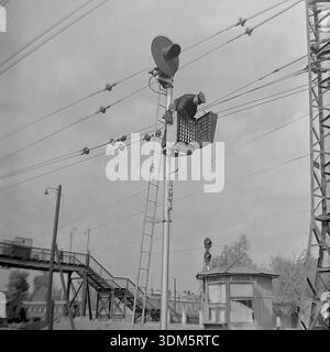 Slaviansk, regione di Donetsk, RSS Ucraina, URSS. 1970 anni Un'autentica vista d'archivio di un segnalatore ferroviario che esegue lavori di manutenzione in quota. Un uomo in abbigliamento da lavoro si trova su un piolo della scala, appoggiato all'alloggiamento aperto della luce segnaletica "ChM3" per riparare la matrice dell'indicatore di percorso. Sotto, sono visibili un ponte pedonale in metallo e un piccolo chiosco della stazione che vende dolciumi. L'immagine cattura il pericoloso lavoro quotidiano del personale tecnico e l'infrastruttura della stazione del pacifico Donbass. Foto Stock