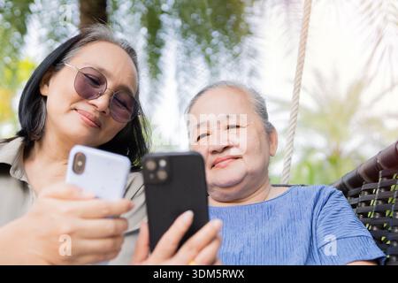 Donna asiatica anziana che esplora la tecnologia, ridendo e guardando il telefono cellulare insieme all'esterno. Vista ad angolo basso, messa a fuoco selettiva. Foto Stock
