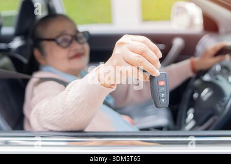 Donna asiatica che sorride e mostra le nuove chiavi della macchina e si siede all'interno della macchina. Vista laterale, messa a fuoco selettiva. Foto Stock