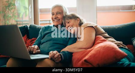 Coppia di anziani felice che si rilassa su un divano a casa mentre utilizza un laptop insieme. Partner maturi sorridenti che apprezzano la vita digitale, la connessione e tutti i giorni Foto Stock