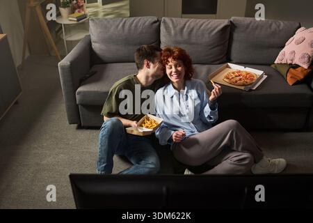 Giovane uomo adulto caucasico seduto al piano accanto alla giovane donna adulta caucasica che mangia pizza e patatine fritte insieme nel soggiorno, sorridendo e gustando il cibo mentre guardi la televisione Foto Stock