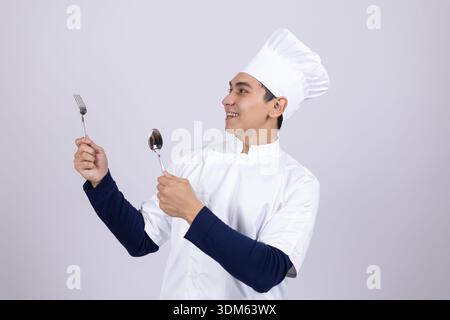 Giovane chef che tiene in mano utensili da cucina e assaggia cibo Foto Stock