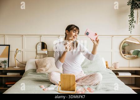 Donna che applica il rullo facciale mentre crea contenuti sullo smartphone in camera da letto Foto Stock