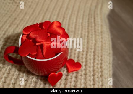 Le decorazioni rosse a forma di cuore riempiono una tazza rossa appoggiata su uno sfondo chiaro e testurizzato. Questa scena trasmette un'atmosfera calda e romantica, perfetta per le celebrazioni di San Valentino. Foto Stock