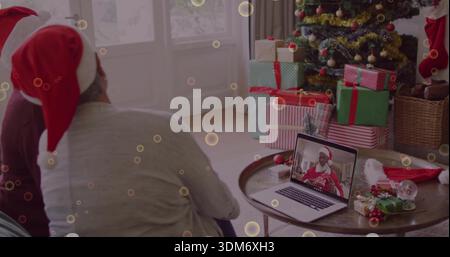 Una coppia seduta che indossa cappelli di Babbo Natale rossi e bianchi a casa, mentre guarda chi chiama il laptop con regali d'albero Foto Stock