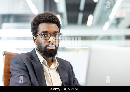 Uomo africano professionista che indossa un abito e occhiali da vista. Guardando direttamente la fotocamera. Trasmettere professionalità e sicurezza aziendale mentre si lavora su un notebook in un ambiente d'ufficio moderno Foto Stock