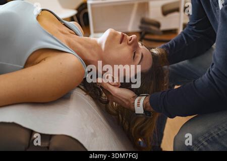 Vista laterale di un fisioterapista professionista che esegue la mobilizzazione del collo per una donna rilassata su un divano medico. Sessione di assistenza sanitaria incentrata sull'osteopa Foto Stock