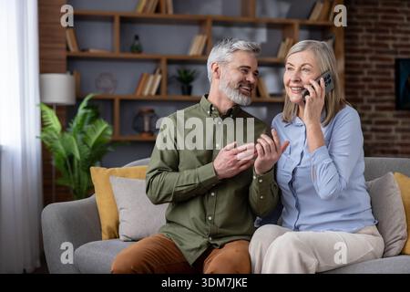 Una coppia matura seduto su un divano nel loro soggiorno. Sorridere e interagire mentre la donna parla attivamente al telefono cellulare. Rappresenta la connessione. Comunicazione. E la felicità in pensione Foto Stock