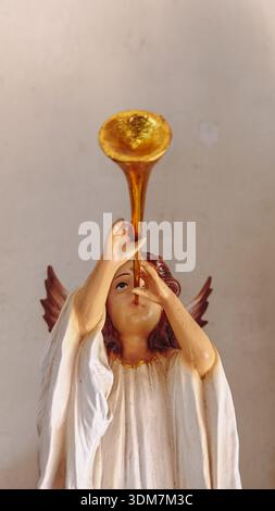 Tromba dorata soffiante con statuina d'angelo, accappatoio bianco, collana d'oro, capelli marroni, ali, sfondo semplice. Foto Stock
