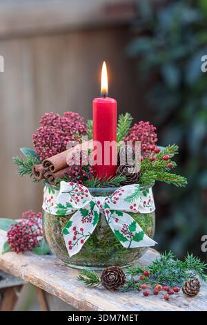 composizione natalizia con candela rossa in un bicchiere decorato con fiori di skimmia, coni, rami di conifere, rosone e involtini di cannella Foto Stock