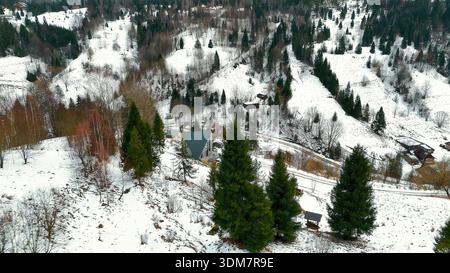 Vista aerea del villaggio di Snowy Mountain. Vista aerea panoramica di un villaggio di montagna innevato con cabine sparse, fitti alberi sempreverdi e strade tortuose Foto Stock