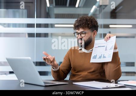 Giovane professionista con capelli ricci e occhiali che partecipa attivamente a una videochiamata, presentando un report finanziario con grafici e grafici mentre lavora in remoto da un moderno ufficio Foto Stock
