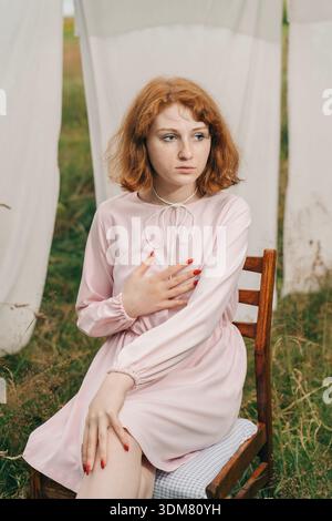 Ritratto di una giovane donna dai capelli rossi in un morbido abito rosa seduta su una sedia in legno vintage in un ambiente naturale all'aperto. Circondato da biancheria appesa Foto Stock