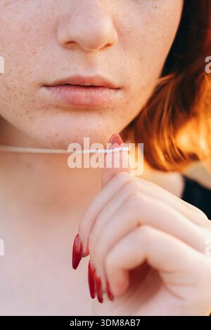 Un primo piano morbido e intimo di una giovane donna dai capelli rossi che tiene una delicata collana di diamanti vicino alle labbra. Le sue lentiggini naturali, le labbra rosa tenue e. Foto Stock