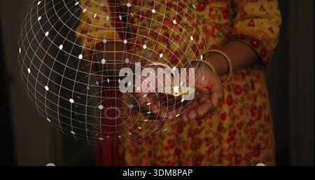 Donna indiana matura che coppetta diya di argilla illuminata in una stanza soffusa, kurta giallo, braccialetti dorati, struttura a reticolo Foto Stock