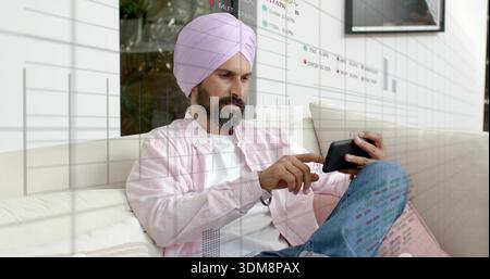 Toccare l'uomo indiano con il turbante rosa e la camicia rosa sul divano, utilizzando lo smartphone con sovrapposizioni finanziarie Foto Stock