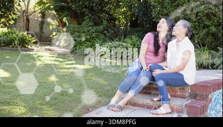 Due donne sedute a sorridere sui gradini della piscina in mattoni nel cortile posteriore, indossando jeans e sandali, spazio per copiare Foto Stock
