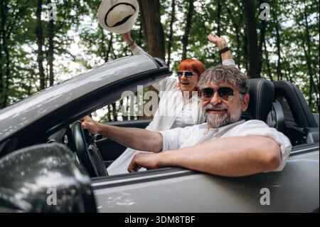 Le mani sono alzate, la donna felice è con il cappello. Coppia senior che guida cabriolet convertibile. Foto Stock