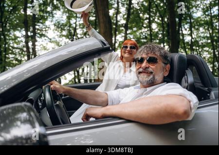 Le mani sono alzate, la donna felice è con il cappello. Coppia senior che guida cabriolet convertibile. Foto Stock