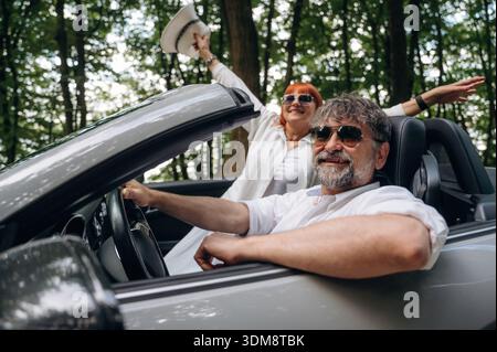 Le mani sono alzate, la donna felice è con il cappello. Coppia senior che guida cabriolet convertibile. Foto Stock