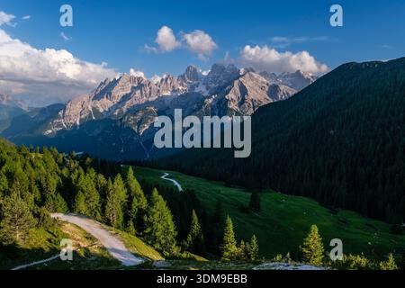 Tortuosa strada di ghiaia attraverso verdi prati alpini. Massicce formazioni rocciose del Monte cristallo in lontananza sotto un cielo blu con morbide nuvole. Foto Stock