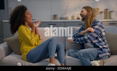 Coppia multietnica uomo donna che parla a casa divano condividere buone notizie eccitato felice insieme maschio caucasico afroamericano femminile stupito gossip Foto Stock
