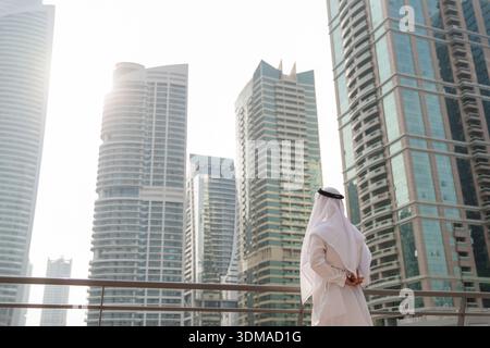 Uomo d'affari degli Emirati che indossa abiti tradizionali bianchi e guarda agli investimenti immobiliari e agli edifici. Foto Stock