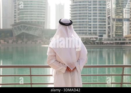 Uomo degli Emirati che indossa abiti tradizionali bianchi in piedi presso la passeggiata di Dubai Marina e guarda il canale. Foto Stock