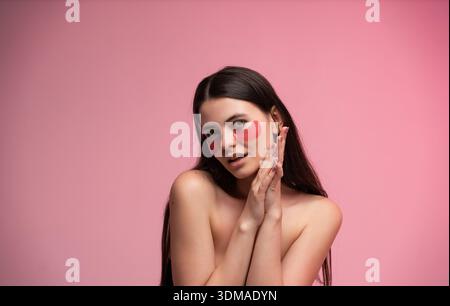 Giovane donna che tocca il viso mentre indossa delle toppe rosse sotto gli occhi durante il trattamento idratante degli occhi su sfondo rosa Foto Stock