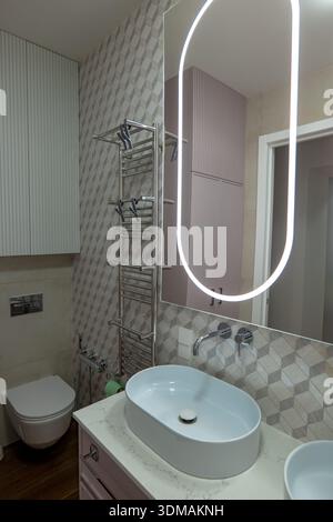 Il bagno contemporaneo presenta un lavandino ovale galleggiante su lavandino in marmo, rubinetto montato a parete, grande specchio con bordo rettangolare illuminato a LED Foto Stock