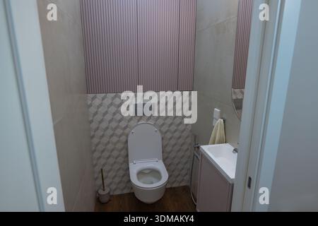 I bagni compatti mostrano WC a parete con coperchio aperto, spruzzi geometrici di piastrelle, armadi a lastre di malva, piccolo lavandino, spruzzatore bidet, piastra a filo, pi Foto Stock