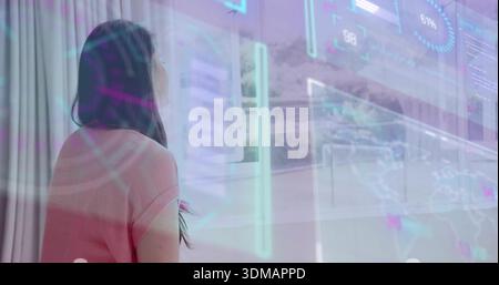 Donna asiatica in piedi con la parte superiore rosa che guarda verso la finestra con sovrapposizioni olografiche dell'interfaccia utente, copia spazio Foto Stock