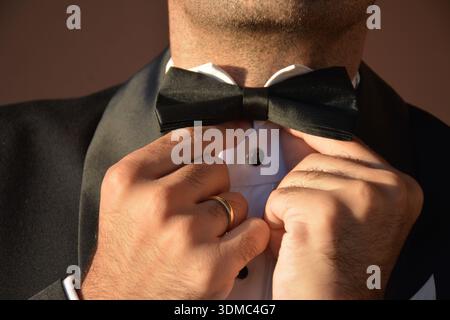 Primo piano delle mani di uno sposo che regola il suo papillon nero. Indossa un abito da smoking nero e una camicia bianca con bottoni neri. Dettaglio delle mani Foto Stock
