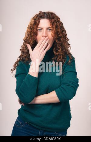Giovane donna dai capelli rossi con un maglione dolcevita verde e jeans copre la bocca con la mano, esprimendo shock o segretezza. Foto Stock