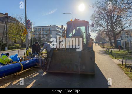 Poznan, Polonia - 18 novembre 2025: Il team di costruzione installa sezioni di condutture blu con una terna gialla, vitale per il servizio urbano continuo Foto Stock