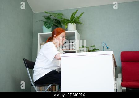 Una vista lontana e con un angolo basso mostra una donna anziana seduta a un tavolo in casa. copia spazio, può essere utilizzato per argomenti relativi alla salute come check-up giornalieri, assistenza agli anziani, o concetti generali di stile di vita. Foto Stock