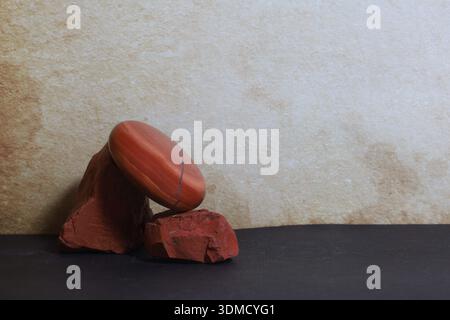 Una pietra rossa lucida di jasper appoggiata su rocce grezze di jasper, posta su uno sfondo neutro dai toni caldi. Foto Stock