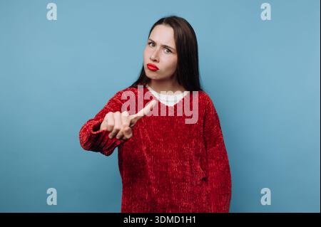 Una persona si trova di fronte a uno sfondo azzurro. Hanno i capelli lunghi e indossano un maglione rosso. La persona alza una mano con uno sguardo serio, signa Foto Stock