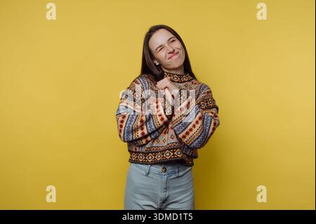 Una giovane donna si trova di fronte a un muro giallo brillante, sorridente e divertente. Indossa un maglione a motivi geometrici e jeans azzurri, mostrando una casu Foto Stock